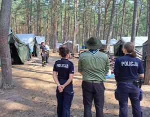 Dwie policjantki i harcerz odwróceni tyłem w tle obóz harcerski rozmieszczony między drzewami.