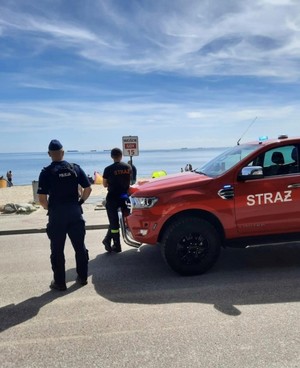 policjant i strażak stoją przy plaży odwróceni tyłem obok wóz strażacki.
