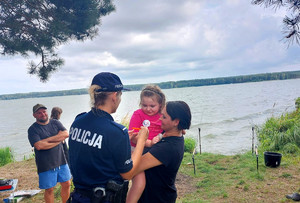 Policjantka podaje odblask dziecku, które na rękach trzyma opiekun w tle dwóch mężczyzn i brzeg zbiornika wodnego.