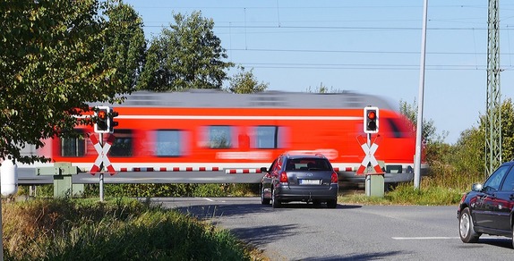 przejazd kolejowy z opuszczonym szlabanem przed którym stoi samochód. Po torach jedzie pociąg