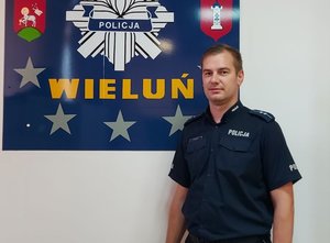 umundurowany policjant pozuje do zdjęcia na tle tablicy przedstawiającej gwiazdę policyjna z napisem Wieluń oraz herby gminy i powiatu wieluńskiego