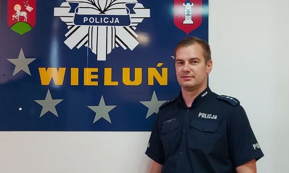 umundurowany policjant pozuje do zdjęcia na tle tablicy przedstawiającej gwiazdę policyjna z napisem Wieluń oraz herby gminy i powiatu wieluńskiego