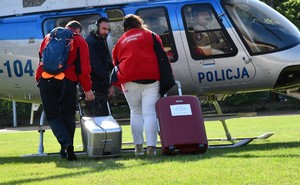 Kobieta i mężczyzna rozmawiają tuż przy policyjnym śmigłowcu stojącym trawiastym lądowisku z mężczyzną ze słuchawkami na uszach.