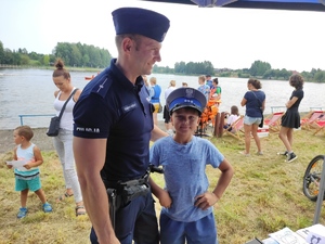 Policjant stoi z obok chłopca, który ma na głowie czapkę policyjną, w tle inni uczestnicy wydarzenia.