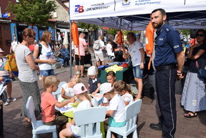 Dzieci siedzą na krzesełkach przy małym stoliku i kolorują obrazki obok stoi policjant w tle uczestnicy imprezy i namiot profilaktyczny Policji.
