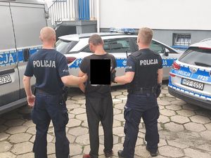 dwaj policjanci z zatrzymanym mężczyzną, w tle zaparkowane radiowozy