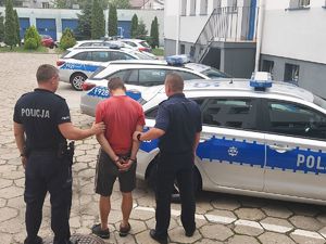 dwaj policjanci z zatrzymanym mężczyzną, w tle zaparkowane radiowozy