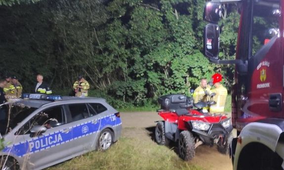 Pora nocna. Z lewej strony widoczny jest policyjny radiowóz i stojący przy nim strażacy, z prawej widać funkcjonariuszy straży przy quadzie, i fragment wozu strażackiego