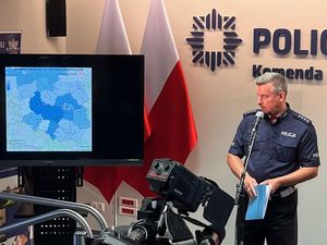 oficer policji w mundurze podczas briefingu na salce konferencyjnej stoi przed mikrofonem i zwraca sie w bok, gdzie na ekranie wyświetlona jest mapa wypadków ze skutkiem śmiertelnym. Z przodu widać kamery. W tle na ścianie kawałek napisu Policja i logo Policji