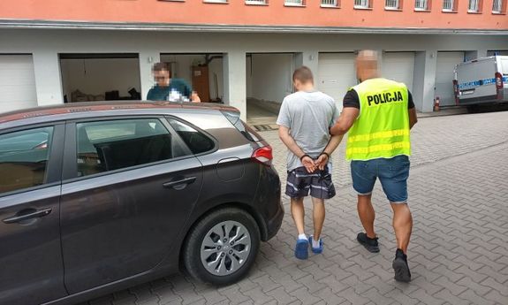 policjant prowadzi zatrzymanego do samochodu. W tel widać budynek, a z boku policyjny radiowóz