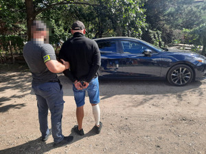 policjant prowadzi zatrzymanego mężczyznę. Policjant ma zasłoniętą twarz, jest ubrany w szarą koszulkę na krótki rękaw i ciemne długie spodnie. Na prawym przedramieniu ma odblaskową opaskę z napisem POLICJA. Po jego prawej stronie stoi zatrzymany mężczyzna. Mężczyzna ten ma czarną czapkę, czarną bluzę i granatowe spodenki o długości do kolan. Prawą łydkę ma obwiązaną bandażem. Na rękach trzymanych z tyłu ma założona kajdanki. W tle widać granatowy pojazd odzyskany w wyniku policyjnej akcji. Obaj mężczyźni stoją przodem do pojazdu i tyłem do obiektywu aparatu.