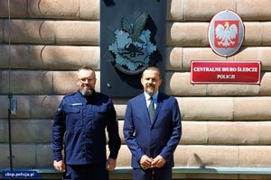 umundurowany policjant i mężczyzna w garniturze pozują do zdjęcia. W tel widoczny jest napis na ścianie Centralne Biuro śledcze Policjii i logo CBŚP