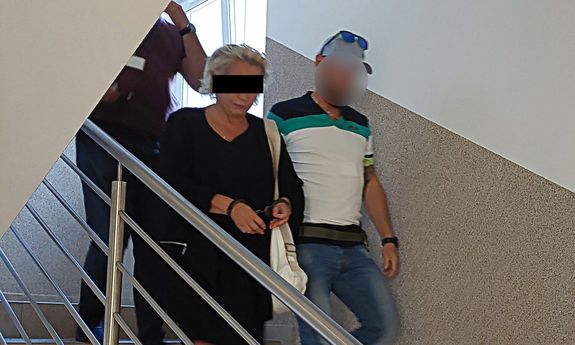 Dwaj policjanci prowadzą zatrzymaną kobietę, która na rękach trzymanych z przodu, ma założone kajdanki