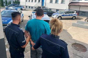 umundurowani policjanci prowadzą zatrzymanego mężczyznę. zdjęcie zrobione na placu wewnętrznym komendy. w tle radiowozy i budynki komendy