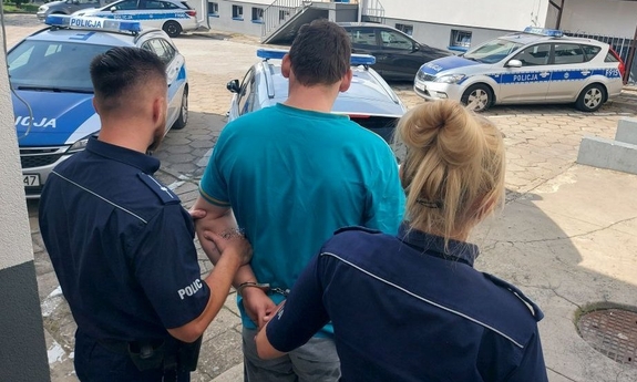 umundurowani policjanci prowadzą zatrzymanego mężczyznę. zdjęcie zrobione na placu wewnętrznym komendy. w tle radiowozy i budynki komendy