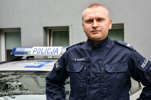 umundurowany policjant pozuje do zdjęcia, w tle widać fragment policyjnego radiowozu i budynek