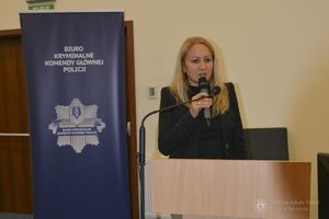 kobieta stoi za mównicą przy mikrofonie, obok niej widoczny baner z napisem Biuro Kryminalne Komendy Głównej Policji