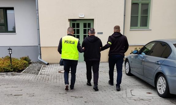 dwaj policjanci prowadzą zatrzymanego do budynku. Policjant z lewej ma na sobie żółtą odblaskową kamizelkę z napisem Policja, Policjant będący po prawej tronie ma na ramieniu odblaskową opaskę z napisem Policja