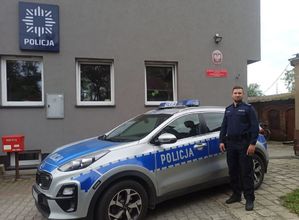 Umundurowany policjant stoi przy radiowozie. W tle widać budynek Policji