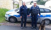przewodnik z psem służbowym, obok drugi policjant, stojący przodem do zdjęcia, za nimi bok radiowozu