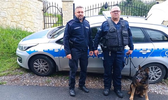 przewodnik z psem służbowym, obok drugi policjant, stojący przodem do zdjęcia, za nimi bok radiowozu