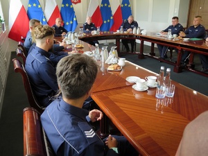 Zdjęcie szczytu owalnego stołu prezydialnego przy którym siedzą nadinsp. Roman Kuster, gen. insp. Jarosław Szymczyk oraz insp. Robert Kumor, w tle godło Polski oraz flagi Polski i UE. Po bokach stołu widoczni siedzący umundurowani policjanci.