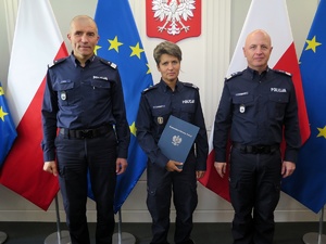 Gen. insp. Jarosław Szymczyk oraz nadinsp. Roman Kuster, pośród których na tle godła i flag: Polski i UE stoi umundurowana policjanta z granatową teczką gratulacyjną.