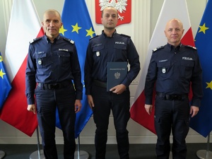 Gen. insp. Jarosław Szymczyk oraz nadinsp. Roman Kuster, pośród których na tle godła i flag: Polski i UE stoi umundurowany policjant z granatową teczką gratulacyjną.