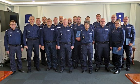 Zdjęcie grupowe 18 umundurowanych policjantek i policjantów na tle bieli ściany, wśród których stoją: gen. insp. Jarosław Szymczyk, nadinsp. Roman Kuster oraz insp. Robert Kumor.