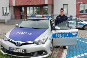 umundurowany policjant stoi przy radiowozie, w tle budynek komendy policji