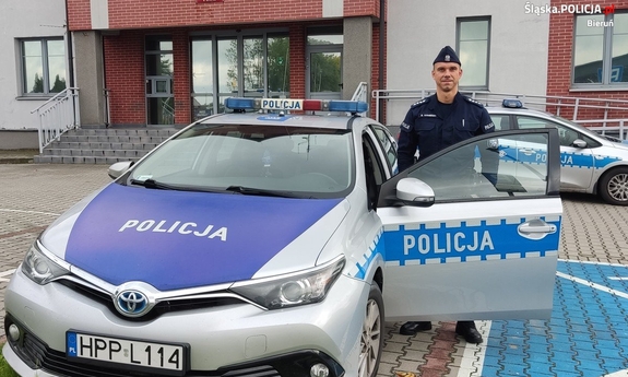 umundurowany policjant stoi przy radiowozie, w tle budynek komendy policji