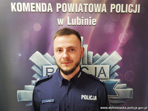 umundurowany policjant - dzielnicowy stoi na tle ścianki z napisem Komenda Powiatowa Policji w Lubinie i gwiazdą policyjną