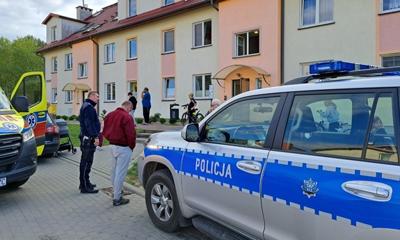na zdjęciu obok radiowozu policyjnego stoi funkcjonariusz i mężczyzna, obok policjanta stoi zaparkowana żółta karetka pogotowia, w tle budynek mieszkalny