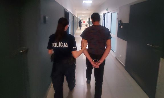 Zatrzymany prowadzony z kajdankami założonymi na ręce trzymane z tyłu. Korytarz komendy. Zatrzymanego prowadzi umundurowana policjantka. Zatrzymany siedzi na krześle w pokoju przesłuchań. Obok stoi umundurowana policjantka