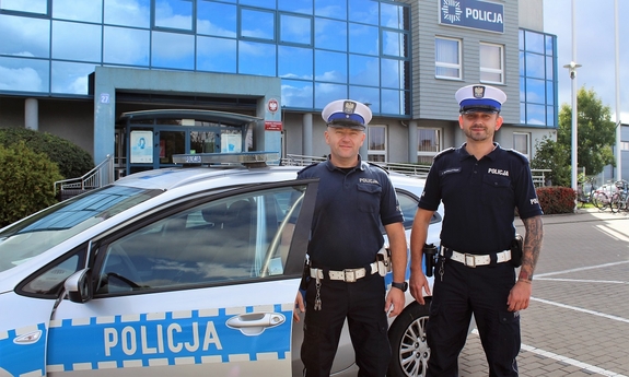 dwóch umundurowanych policjantów stoi przy radiowozie, w tle budynek komendy policji