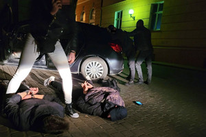 dwóch mężczyzn leży brzuchem do ziemi. Nad nimi pochyla się nieumundurowana policjantka i trzyma ich za odzież. Za nimi stoi otwarty samochód. Na końcu auta policjant trzyma trzecią osobę zatrzymaną