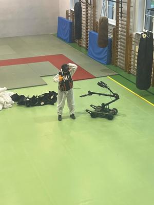 Sala gimnastyczna - widok z góry  z pewnej odległości. Człowiek ubrany na biało  w czarnym kasku, w kamizelce na której ma ładunki wybuchowe. Przy nim widoczny jest robot do rozbrajania ładunków wybuchowych.