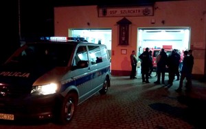 Pora nocna. grupa funkcjonariuszy straży i policji przed jednostką Ochotniczej Straży Pożarnej. W tle w garażu widać wóz straży pożarnej, przed budynkiem stoi radiowóz