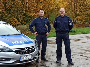Dwaj policjanci stojący obok radiowozu w terenie zielonym