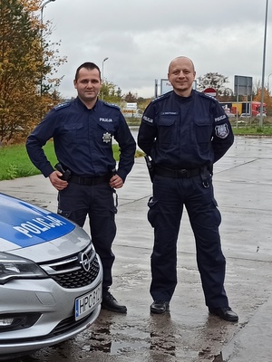 Dwaj policjanci stojący obok radiowozu w terenie zielonym
