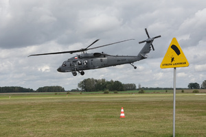 Policyjny śmigłowiec Black Hawk nisko nad trawiastym lądowiskiem, z prawej żółty znak z sylwetką spadochroniarza i napisem strefa lądowań.