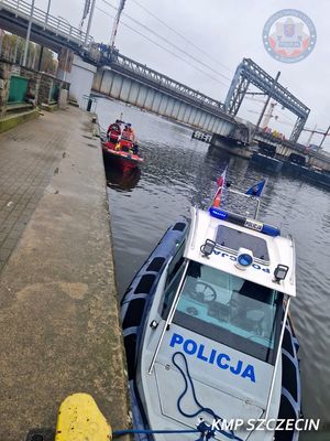 widok z nabrzeża, na pierwszym planie policyjna motorówka, z tyłu zacumowana przy nabrzeżu łódź WOPR a na niej dwaj ratownicy i mężczyzna owinięty kocem termicznym