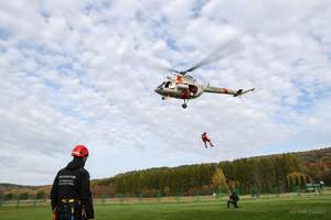 śmigłowiec w powietrzu i jeden z funkcjonariuszy na linie podwieszonej pod śmigłowiec, drugi trzyma końcówkę liny. z boku stoi instruktor ratownictwa wysokościowego