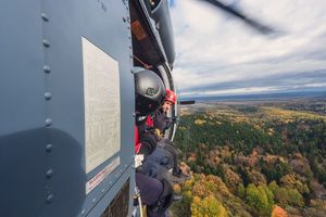 funkcjonariusze siedzą na brzegu śmigłowca w locie i patrzą na okolicę nad którą przelatują