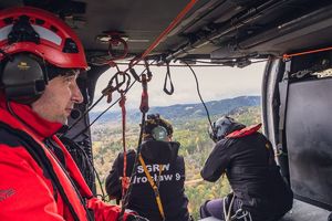 dwaj funkcjonariusze siedzą na brzegu śmigłowca w locie patrzą w dół