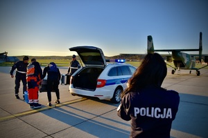 mężczyźni stoją na płycie lotniska i wyciągają z bagażnika pojazdu walizkę, obok nich stoi umundurowana policjantka, w tle śmigłowiec gotowy do lotu