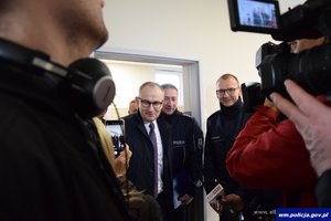 na pierwszym planie część twarzy policjanta i kamerzysty, który nagrywa mężczyznę w garniturze i stojących wokół niego oficerów Policji