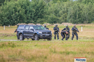 Policyjni kontrterroryści prowadzą zatrzymanego polną drogą, za nimi samochód typu SUV.