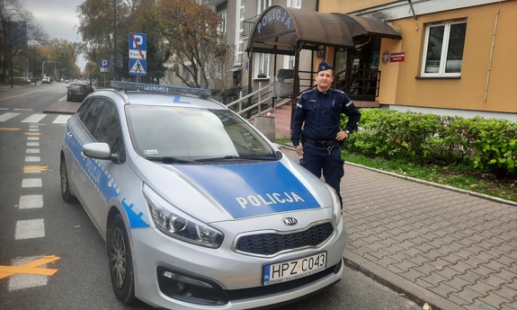 umundurowany policjant stoi przy radiowozie, w tle budynek jednostki policji