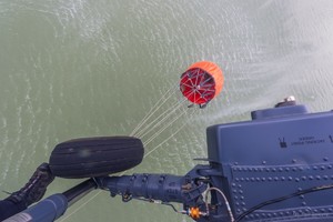 Czerwony pojemnik Bambi Bucket podwieszony pod policyjnym śmigłowcem Black Hawk nad zbiornikiem wodnym.